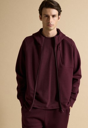 Joven con sudadera con capucha burdeos oscura con cremallera, camiseta a juego y pantalones, de pie contra un fondo beige liso.