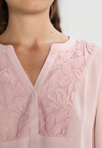 Blouse rose avec un panneau en dentelle florale texturée sur l'empiècement, col en V et tissu doux et lisse.