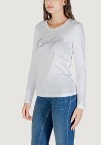 T-shirt bianca a maniche lunghe con scollatura tonda e un design brillante "Live" sul davanti. Abbinata a jeans blu. Materiale in cotone morbido.