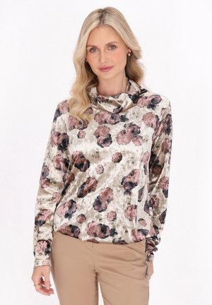 Femeie blondă purtând un top de catifea cu mâneci lungi cu flori roz și negre și pantaloni bej pe fundal alb.