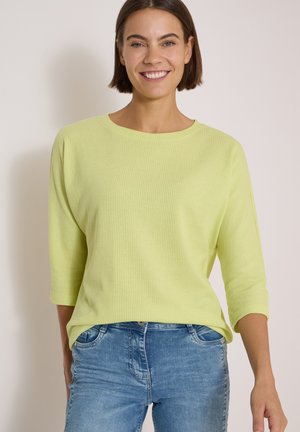 Frau mit kurzen braunen Haaren lächelnd, trägt ein hellgrünes Langarmshirt und blaue Jeans vor einem schlichten hellen Hintergrund.
