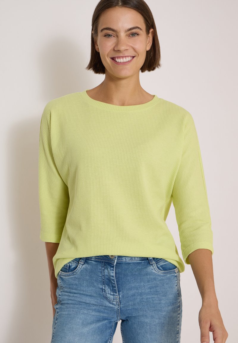 Woman with short brown hair smiling, wearing a light green long-sleeve shirt and blue jeans against a plain light background.