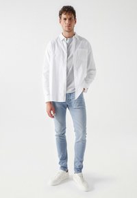Camisa blanca de botones sobre una camiseta gris, combinada con vaqueros ajustados de color azul claro y zapatillas blancas. Se puede ver la tela suave y el corte entallado.