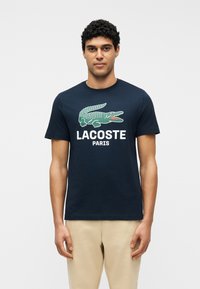 Άνδρας φορώντας μπλε ναυτικό T-shirt Lacoste με μεγάλο πράσινο κροκόδειλο και κείμενο «LACOSTE PARIS», συνδυασμένη με μπεζ παντελόνι, στέκεται μπροστά σε λευκό φόντο.