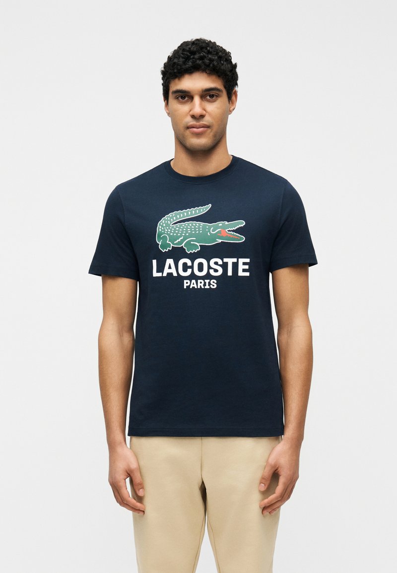 Άνδρας φορώντας μπλε ναυτικό T-shirt Lacoste με μεγάλο πράσινο κροκόδειλο και κείμενο «LACOSTE PARIS», συνδυασμένη με μπεζ παντελόνι, στέκεται μπροστά σε λευκό φόντο.