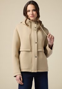 Vaalean beige huppari, jossa on napit, säädettävä naru ja etutaskut. Valmistettu pehmeästä kankaasta, säännöllinen istuvuus ja sileä pinta.