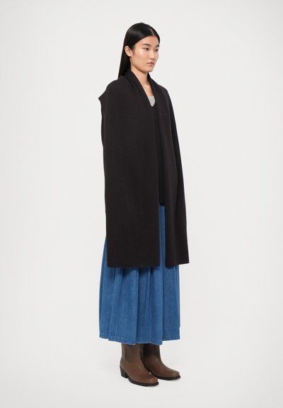 Pull en laine noir à manches longues et fluides porté sur un haut gris et une jupe en denim bleue, associé à des bottines en cuir marron.