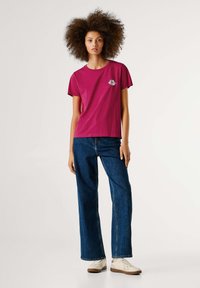 Fuksiškos trumpomis rankovėmis marškiniai su lūpų grafika; derinami su aukštu juosmeniu džinsais; modelis dėvi baltus sportbačius, stovintis prieš šviesų foną.