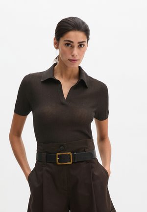 Femme aux cheveux foncés attachés en queue de cheval basse, portant un polo marron foncé ajusté à manches courtes, rentré dans un pantalon marron foncé taille haute avec une ceinture noire.