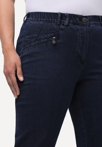 Mörkblå denimbyxor med en slät yta, elastisk midja och en sidoficka med dragkedja. Har en metallknapp som accent.