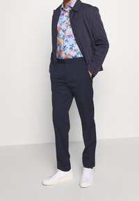 Chaqueta liviana azul marino, camisa de estampado floral en tonos azules y morados, pantalones azul marino ajustados y zapatillas blancas. Estética limpia y moderna.
