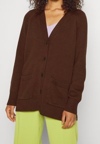 Hnědý pletený cardigan s výstřihem ve tvaru V, dlouhými rukávy, zapínáním na knoflíky a dvěma předními kapsami. Látka má měkký, texturovaný povrch.