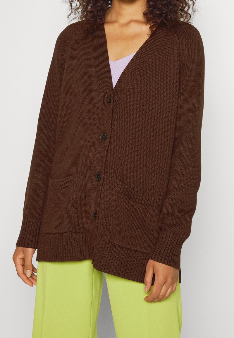 Hnědý pletený cardigan s výstřihem ve tvaru V, dlouhými rukávy, zapínáním na knoflíky a dvěma předními kapsami. Látka má měkký, texturovaný povrch.