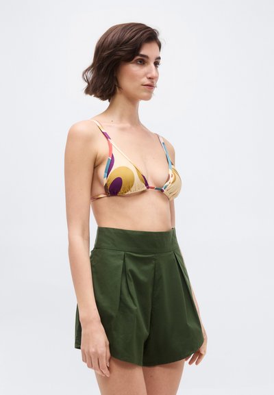 Kvinde iført en bikini top med flerfarvet abstrakt print kombineret med højtaljede, mørkegrønne plisserede shorts, stående sideværts.