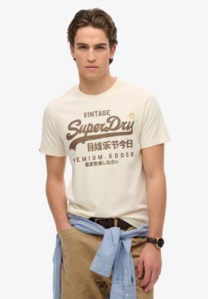 Cream cotton T-shirt featuring a vintage brown "SuperDry" logo graphic. Worn with khaki pants and a light blue shirt tied at the waist.