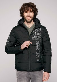 La chaqueta acolchada negra presenta un cierre de cremallera, capucha y gráficos impresos en la parte frontal. El material es suave y ligeramente brillante.