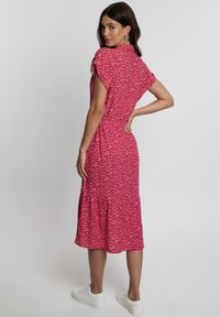 Rotes Kleid mit kurzen Ärmeln, featuring ein pinkfarbenes abstraktes Muster, einem gestuften Saum und einer taillierten Taille, kombiniert mit weißen Sneakers.