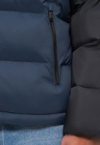 Námornícka puffová bunda s oddielom na zips. Materiál je hladký s matným povrchom. Priľahlá tkanina je čierna a textúrovaná.