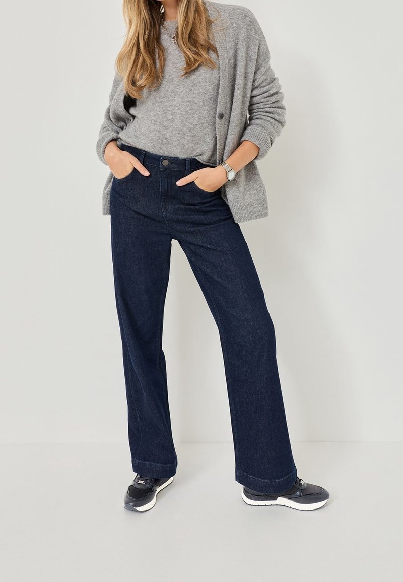 Femme portant un pull gris et un cardigan, jean large foncé, baskets noires, debout les mains dans les poches sur un fond uni.