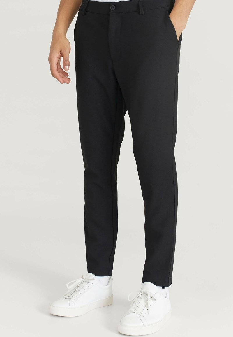 Pantaloni neri slim-fit realizzati in tessuto liscio con chiusura a bottone, dotati di tasche laterali e dettagli di cucitura discreti, abbinati a sneakers bianche.