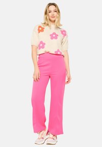 Pull crème avec de grands motifs floraux roses et orange, associé à un pantalon taille haute, large et rose vif, et des chaussures de sport.