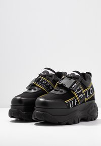 Svarta sneakers med meshstoff och glansiga detaljer, med gula ränder och text, kraftig sula och spännband.