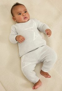 Hallatud hallist kahest osast koosnev rõivakomplekt imikutele, mis sisaldab valge geomeetrilise mustriga kampsunit ja sobivaid pükse, seatud heledate tekstuuriga taustale.