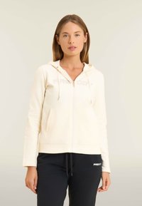 Donna con una felpa cremoso con zip e cappuccio, indossata con pantaloni neri con coulisse, in piedi contro uno sfondo chiaro.
