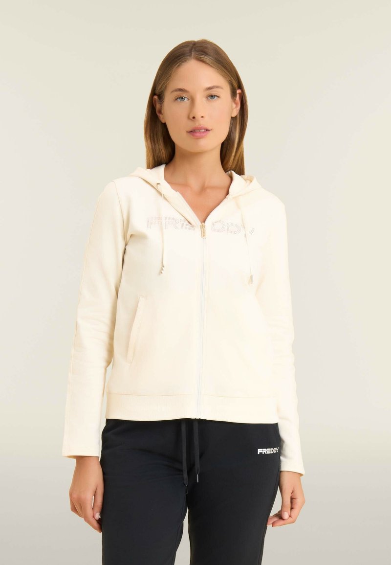 Donna con una felpa cremoso con zip e cappuccio, indossata con pantaloni neri con coulisse, in piedi contro uno sfondo chiaro.
