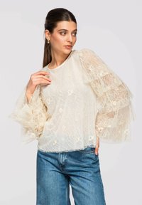 Jeune femme aux cheveux foncés portant un chemisier en dentelle crème à manches cloche superposées et un jean bleu, posant devant un fond clair uni.