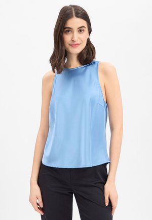 Junge Frau mit schulterlangem dunklem Haar trägt ein ärmelloses hellblaues Satintop und schwarze Hose, steht vor weißem Hintergrund.