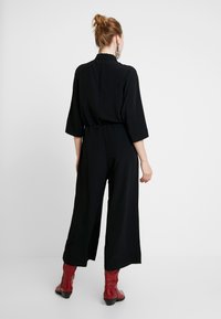 Černý overal s širokými nohavicemi, kimono rukávy a přiléhavým pasem. Nosený s červenými kozačkami po kolena. Pohled zezadu na jednobarevném pozadí.