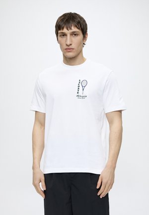 Jeune homme portant un T-shirt blanc Lacoste avec un motif de raquette de tennis et un texte, associé à un short noir, debout devant un fond uni.