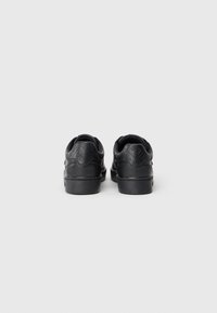 Un paio di sneakers nere viste da dietro con un motivo a logo embossato sul tallone e una suola spessa su uno sfondo bianco.