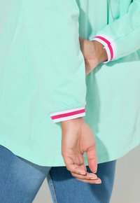 Persona in maglietta a maniche lunghe color turchese chiaro con polsini a righe rosa e bianche, mani dietro la schiena, che indossa jeans blu.