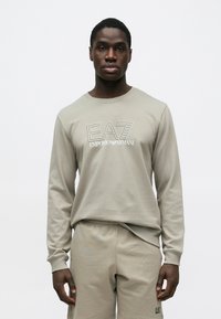 Fiatal férfi, beige Emporio Armani EA7 pulóverben és hozzáillő rövidnadrágban, egyszerű világosszürke háttér előtt áll.