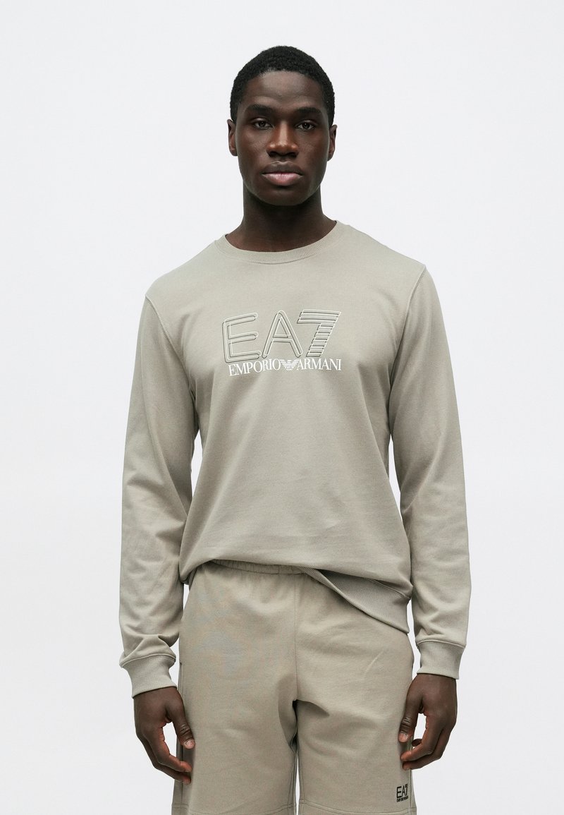 Fiatal férfi, beige Emporio Armani EA7 pulóverben és hozzáillő rövidnadrágban, egyszerű világosszürke háttér előtt áll.