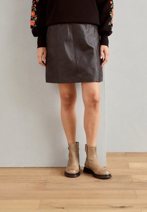 Botas de tobillo de cuero marrón con un acabado suave y tacón bajo, combinadas con una falda mini de cuero negra. Fondo de suelo de madera.
