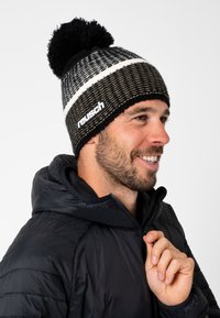 Knitted beanie in black and beige with a pom-pom on top, featuring horizontal stripes and "reusch" logo in white. Worn with a black jacket.