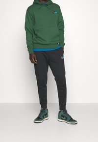 Grön hoodie med känguruficka, blå inre lager, svarta joggers med logotyp, och grön/svarta hög-top sneakers. Avslappnad atletisk outfit.