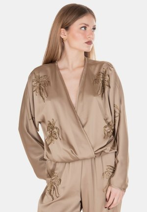 Donna con i capelli lunghi che indossa un outfit in raso beige decorato con disegni di palme ricoperte di perline dorate, guarda verso destra.