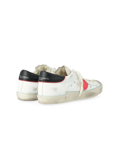 Sneaker bianche basse con tasselli neri sul tallone, toppe circolari rosse sui lati, suole e lati usurati e leggermente sporchi, viste dalla parte posteriore sinistra.