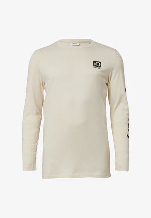 Beige blouse met lange mouwen en een klein zwart Discovery-logo op de borst, en een zwart tekstontwerp dat langs de linker mouw loopt.