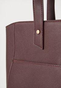 Burgundska torba od glatkog sintetičkog materijala, s zlatnim detaljima i ojačanim šavovima na ručkama i telu.