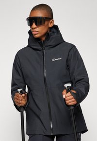 Black waterproof jacket with a hood and front zipper, featuring a logo on the chest. Accessory: black ski poles, user wearing dark oversized sunglasses.