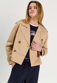 Chaqueta beige de doble pecho con grandes botones, amplios solapas con muesca y longitud corta. Llevada sobre una blusa azul marino con detalles gráficos.