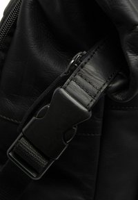 Black leather bag corner featuring a sturdy plastic buckle and strap, with visible stitching and a zipper pull for secure closure.