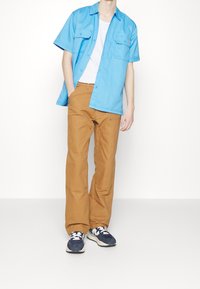 Blauw short-sleeve shirt met twee borstzakken, gecombineerd met tan wijde broek en marineblauwe sneakers. Het shirt heeft een losse pasvorm.