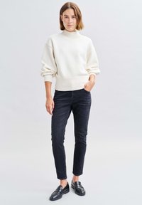 Cremiger, übergroßer Strickpullover mit hohem Kragen und ribbeligem Saum, kombiniert mit enganliegenden schwarzen Jeans und schwarzen Slippern. Glatte Textur.