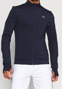 Marin zip-up jacka i stretchig tyg med hög krage, tumhål och Lacoste-logotyp på bröstet. Har sidofickor och kontrasterande kant.